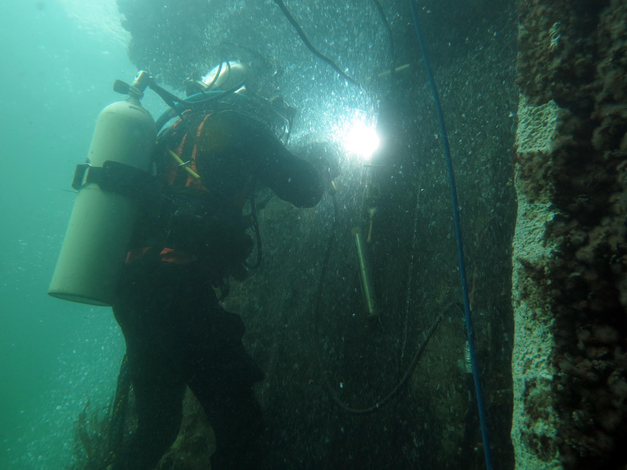 Taucher beleuchtet oder schweißt unter Wasser an einer Wand. Rechts hängen Kabel herab.