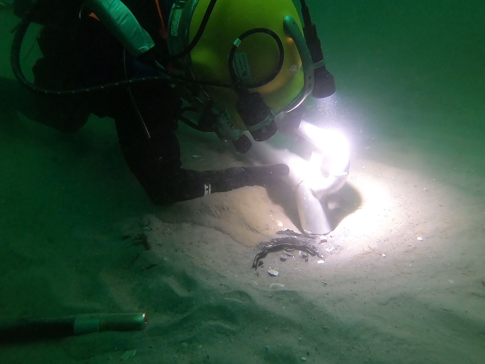 Ein Taucher am Meeresboden beleuchtet metallische Objekte, das Wasser schimmert grünlich