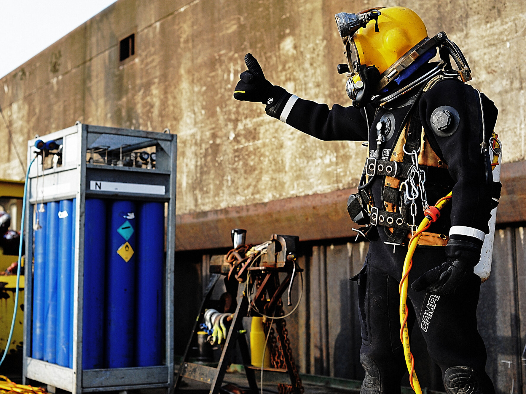 A diver in black diving suit with yellow helmet showing thumbs up.